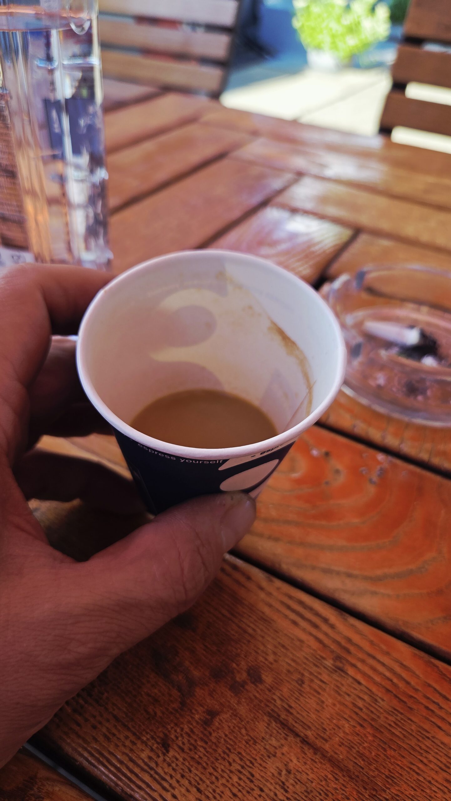A hand holding a small paper cup, half full of coffee, over a wooden table